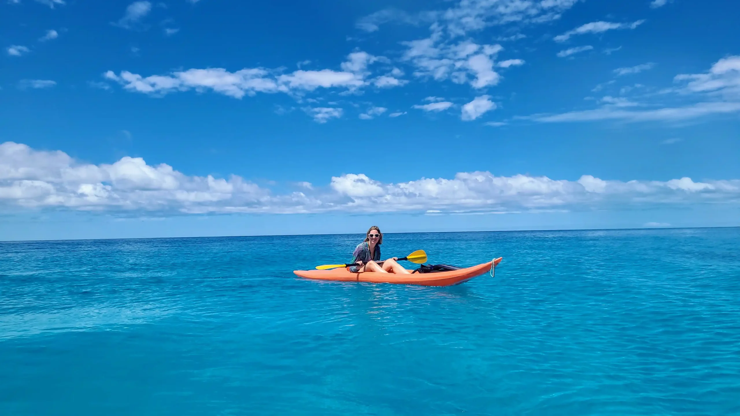 A woman kayaking on a calm, turquoise ocean under a clear blue sky with scattered clouds.