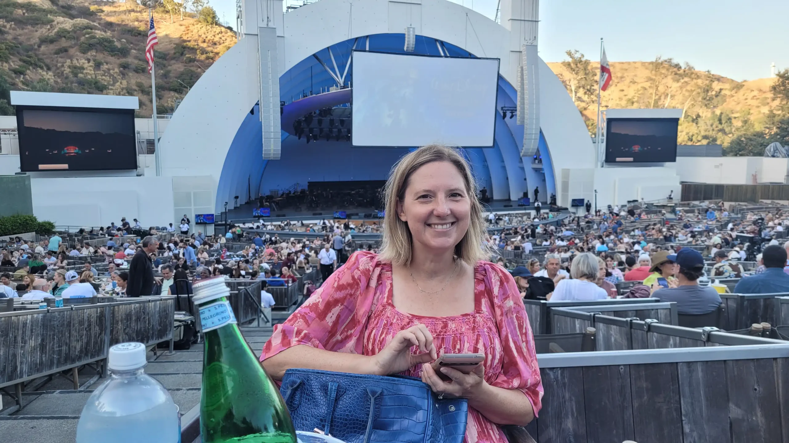 A woman smiling and holding her phone, sitting in an outdoor concert venue with a large crowd and stage in the background.