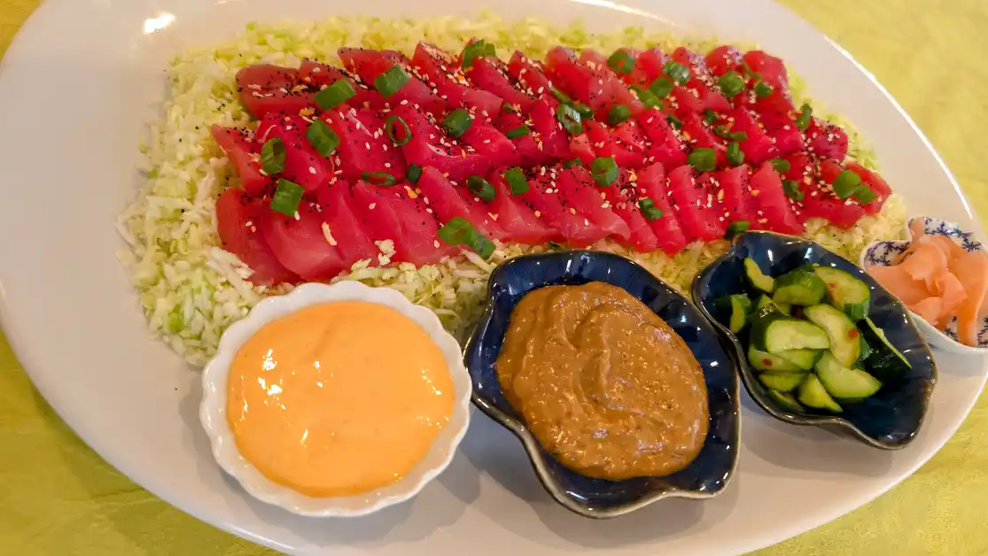 A plate of Tahitian sashimi with sliced Ahi tuna on a bed of shredded cabbage, served with spicy brown sauce, creamy orange sauce, and cucumber salad.