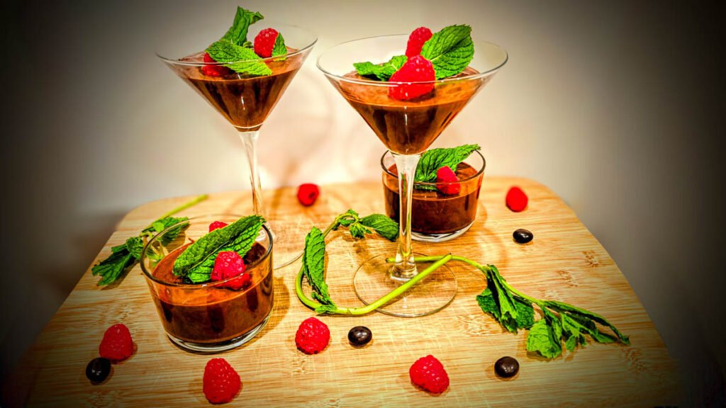 Chocolate mousse desserts garnished with fresh raspberries and mint leaves in various glass containers on a wooden board.