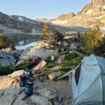 A green tent and camping gear set up on a rocky lakeshore with a majestic mountain backdrop.