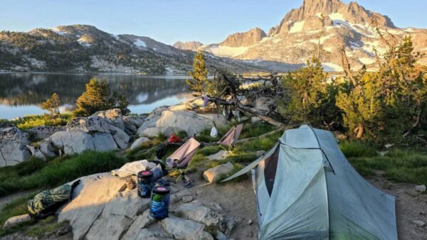 A green tent and camping gear set up on a rocky lakeshore with a majestic mountain backdrop.